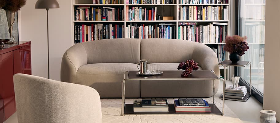 Wohnzimmer mit rundem Sofa vor Bücherwand, Couchtisch mit Tablett und Trauben, Sessel im Vordergrund