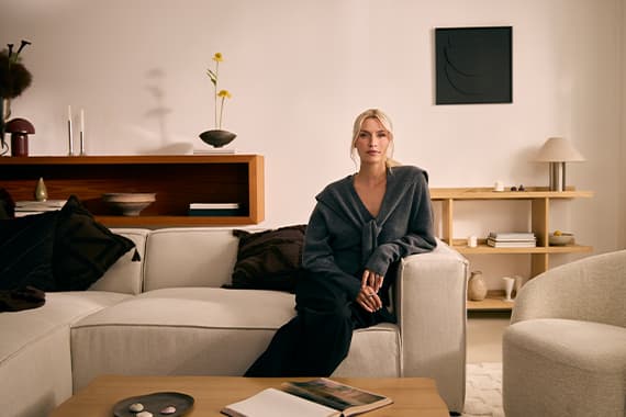 Frau im grauen Cardigan sitzt auf beigem Sofa in modernem Wohnzimmer mit Holzregalen und Deko im Hintergrund