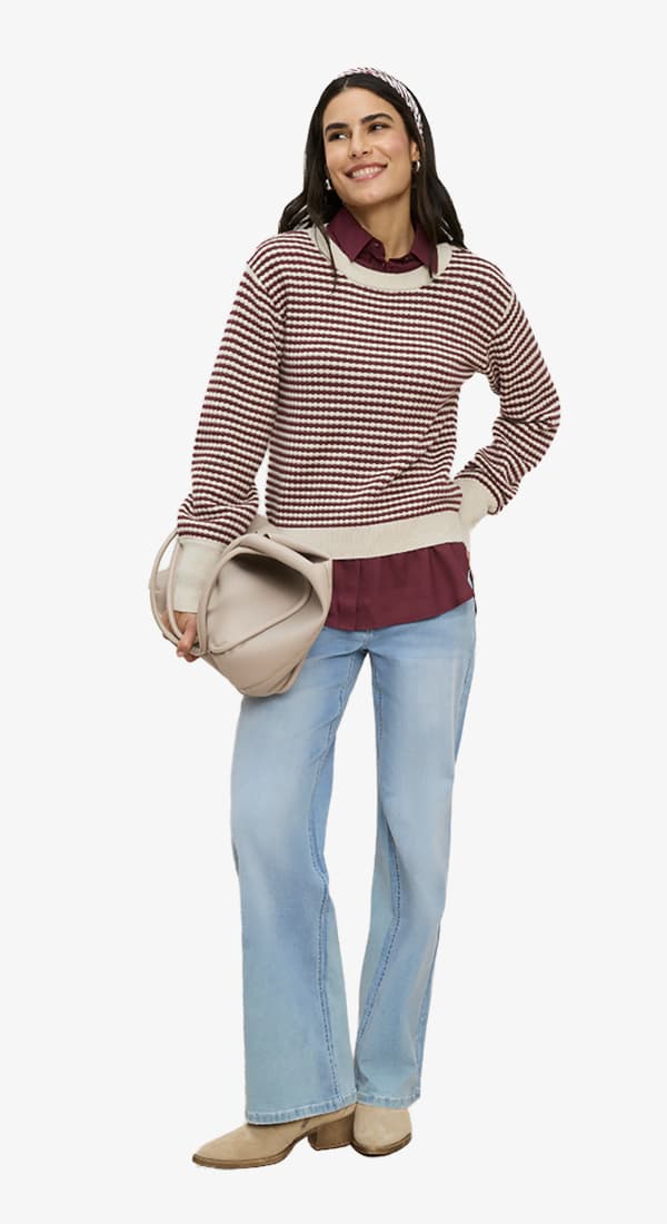 Frau in gemustertem Pullover, Bluse und heller Schlagjeans, hält eine runde Tasche und blickt lächelnd zur Seite