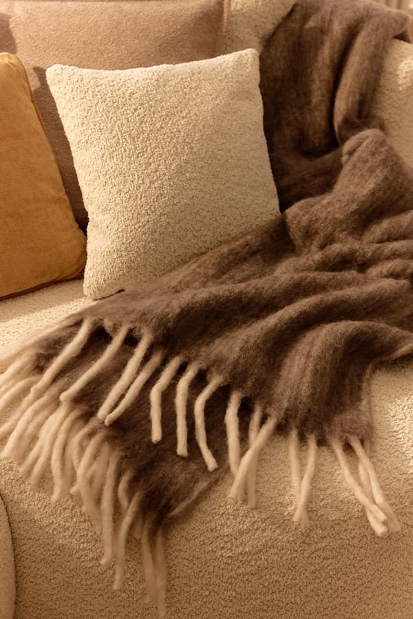 Braune Decke mit langen Fransen liegt locker über cremefarbenem Sofa mit mehreren beigefarbenen und ockerfarbenen Kissen