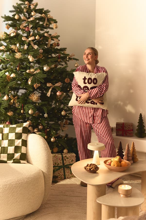 Kissen mit Schriftzug „too tired“ – Frau in gestreifter Pyjama-Hose steht lächelnd vor geschmücktem Weihnachtsbaum