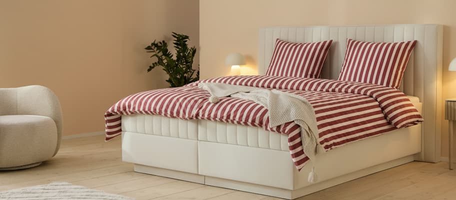 Doppelbett mit rot-weiß gestreifter Bettwäsche, zwei Kissen und Decke, daneben Sessel, Lampen und Pflanze im Schlafzimmer