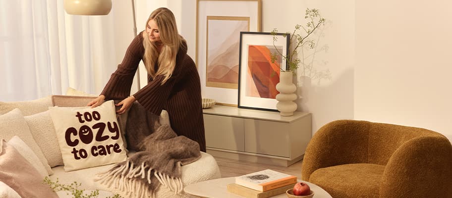 Kissen mit Aufdruck „too cozy to care“ – Frau richtet Sofa in warmem Wohnzimmer mit Bildern und Sessel ein