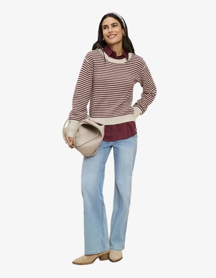 Frau in karierter Strickpullover-Schicht, weiter Jeans und Stiefeletten, hält eine Tasche und schaut seitlich nach vorn