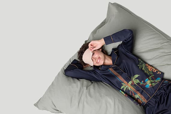 Frau in gemusterter Schlafanzughose liegt auf großem Kissen und schiebt eine Schlafmaske von den Augen