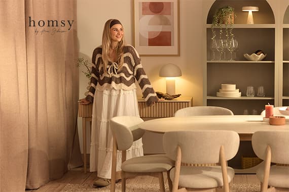 homsy Logo links – Frau im langen Kleid steht lächelnd in warm beleuchtetem Esszimmer mit Tisch und Regal