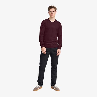Mann steht frontal zur Kamera in dunkelrotem Pullover und dunkler Hose vor hellem Hintergrund