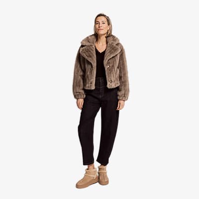 Frau steht lächelnd in brauner Jacke, schwarzer Hose und beigen Stiefeletten vor hellem Hintergrund