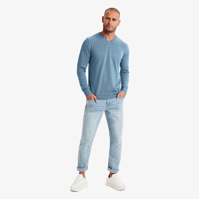 Mann steht frontal, trägt hellblauen Pullover mit V-Ausschnitt, hellblaue Jeans und weiße Sneaker vor hellem Hintergrund