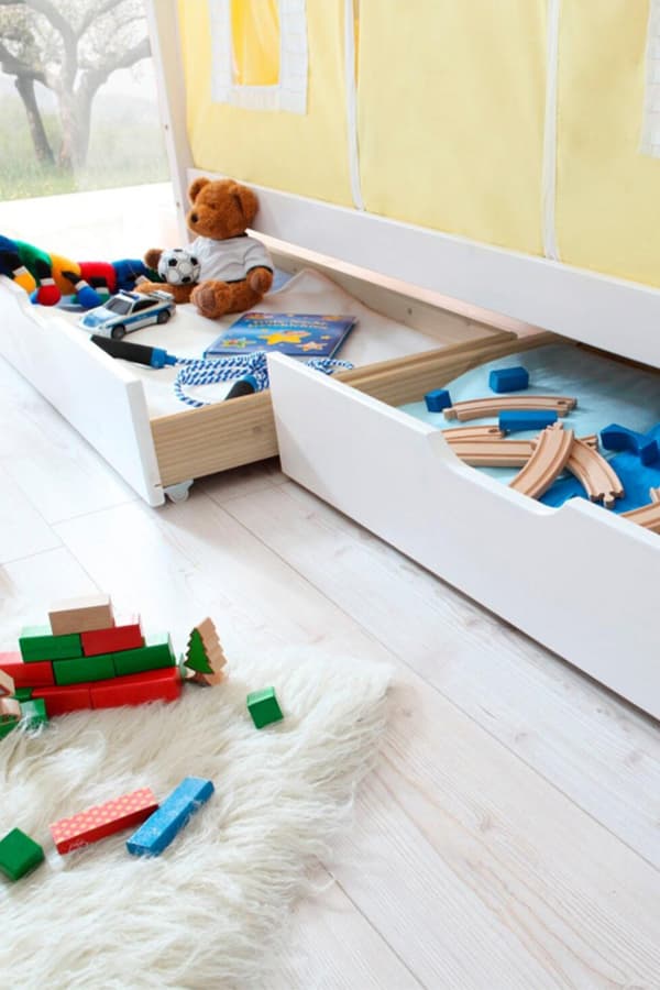 Kinderspielzeug, darunter ein Stoffbär, Holzklötze und Spielzeugautos, in und vor zwei Schubladen unter einem gelben Hochbett