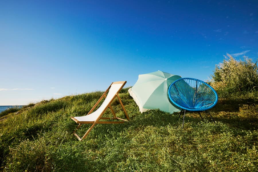 Liegestuhl, blauer Stuhl und hellgrüner Sonnenschirm auf grünem Hügel unter blauem Himmel mit Meer im Hintergrund