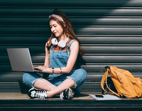 Frau in Jeans-Latzhose sitzt im Schneidersitz, arbeitet am Laptop, gelber Rucksack und Kopfhörer neben ihr auf dem Boden