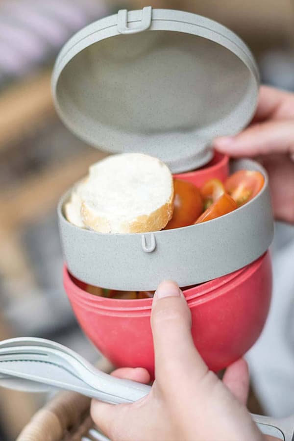 Hand hält grauen Behälter mit Deckel, gefüllt mit Brot und Tomaten, roter Unterteil und Besteck in der anderen Hand