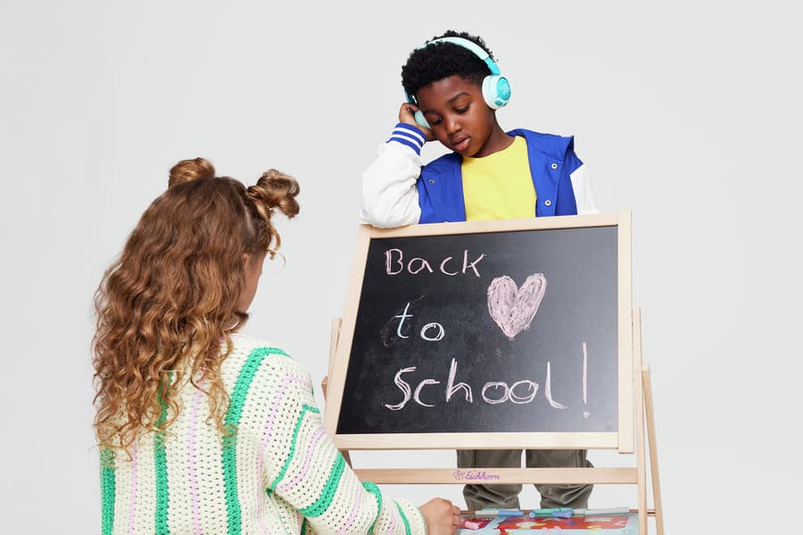 Back to school! – Junge mit Kopfhörern steht hinter Tafel, Mädchen mit lockigem Haar sitzt davor und schaut darauf