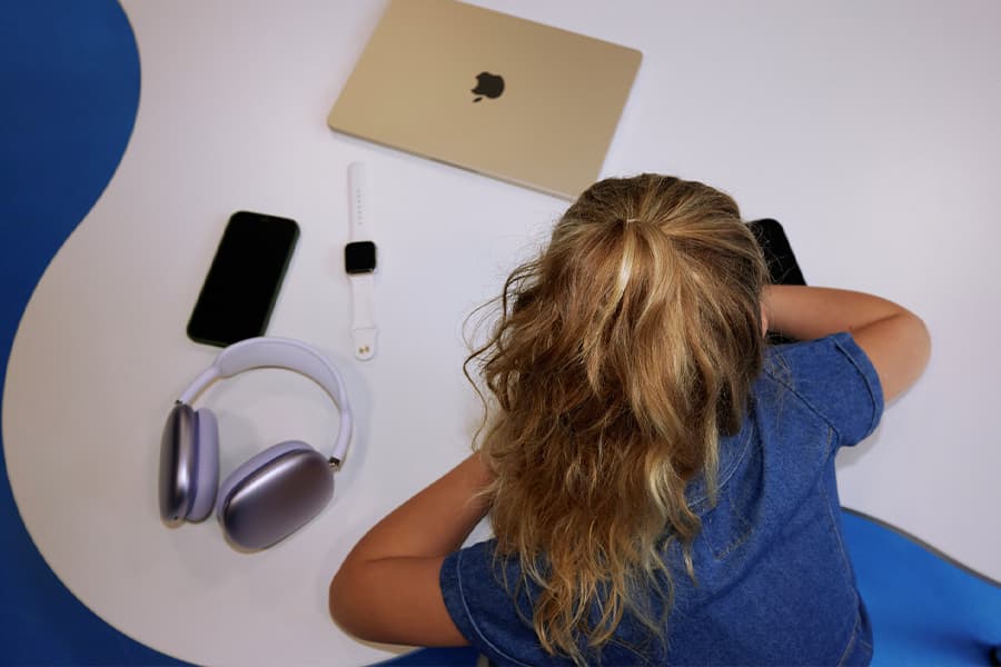 Mädchen mit blonden Haaren sitzt vor einem Tisch mit Laptop, Smartphone, Smartwatch und Kopfhörern.