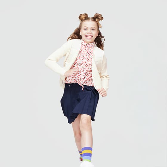 Mädchen in heller Strickjacke, gepunkteter Bluse und blauem Rock läuft, trägt bunte Socken mit Streifen.