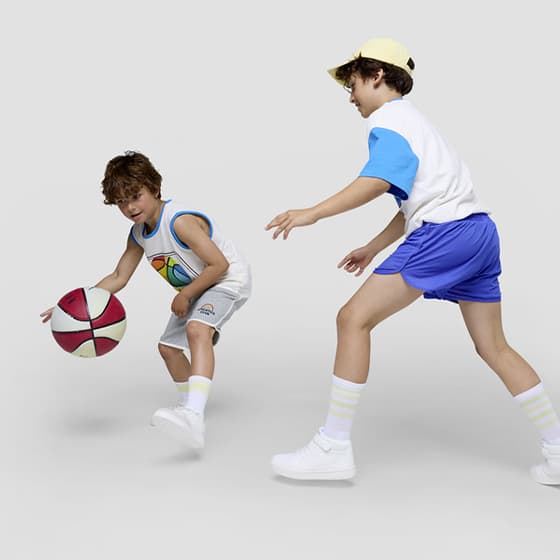 Zwei Kinder spielen Basketball, eines dribbelt den Ball, das andere verteidigt. Beide tragen Sportkleidung und Sneakers.