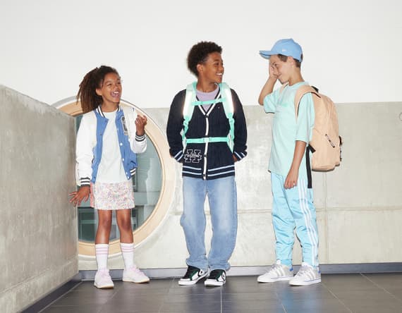 Drei Kinder in Freizeitkleidung stehen nebeneinander, zwei mit Rucksäcken, eines in Jeans und Sneakers, andere in Shorts und Rock.