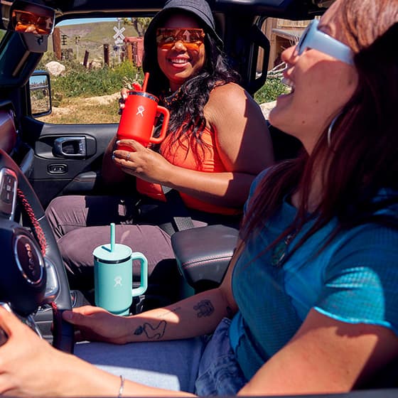 Zwei Personen im Auto, eine hält einen roten Becher mit Strohhalm, die andere hat einen blauen Becher auf dem Armaturenbrett