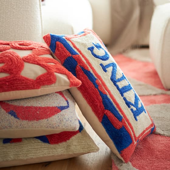 HELLO LOVELY – Rosa Kissen mit gelben Pompons auf weißem Sofa, dahinter weiteres Kissen mit buntem Design