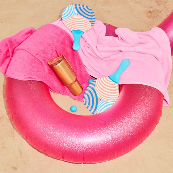 Strandset mit pinkem Schwimmring, zwei bunte Schläger, pinkes Handtuch und orangefarbene Trinkflasche auf Sand.