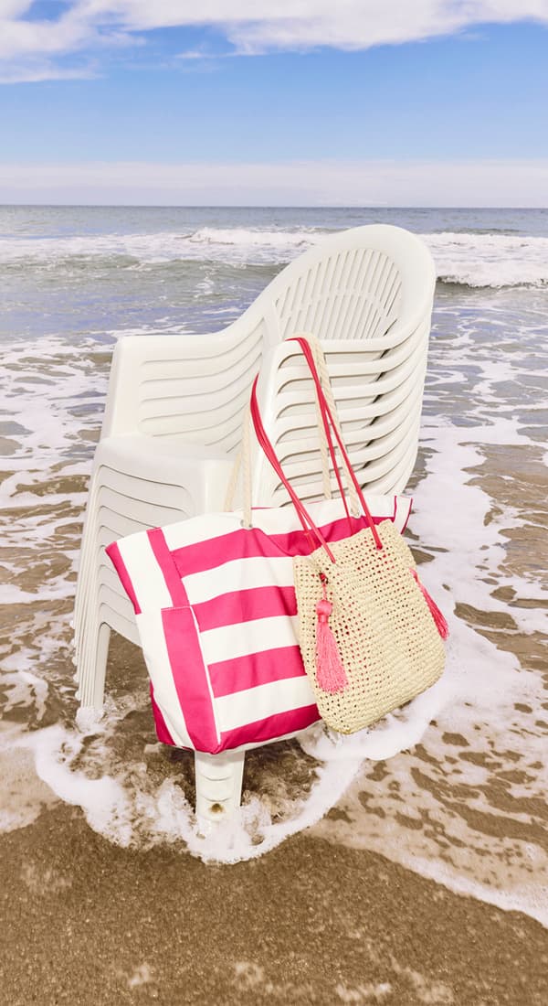 Mehrere weiße Plastikstühle, gestapelt, am Strand. Große gestreifte Tasche und geflochtene Tasche mit Quasten lehnen daran.