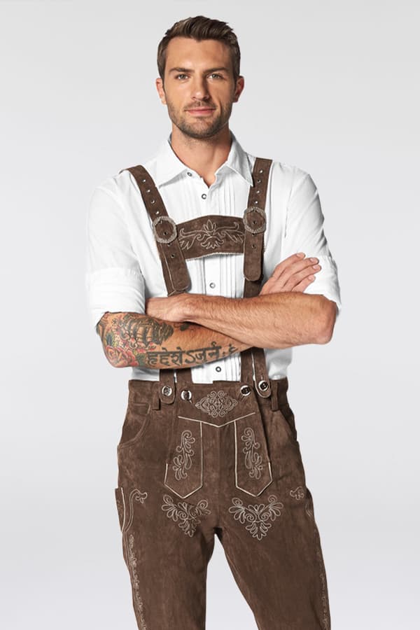 Mann mit tätowierten Armen trägt weiße Trachtenbluse und braune Lederhose mit Stickereien.