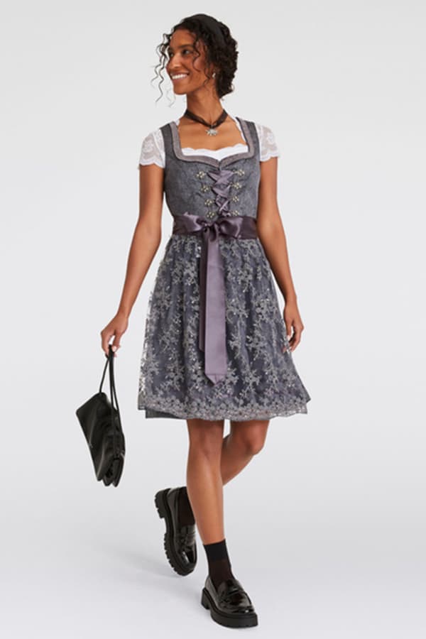 Frau trägt graues Dirndl mit lila Schürze und Spitze, hält schwarze Tasche in der Hand, trägt schwarze Schuhe und Socken