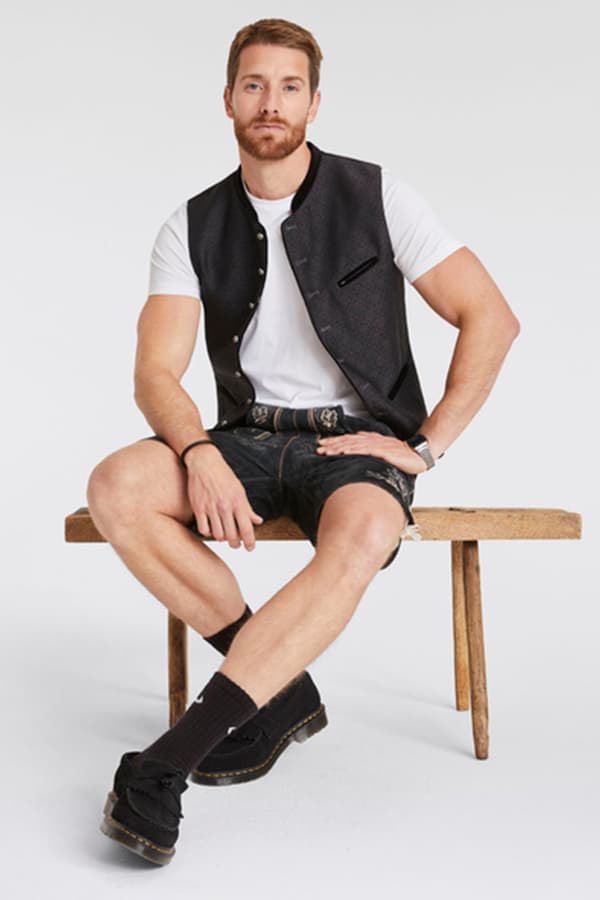 Mann sitzt auf Holzbank und trägt weiße T-Shirt, schwarze Weste, Lederhose und schwarze Schuhe mit Socken