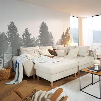 Ecksofa mit Kissen und Decken in hellem Wohnzimmer mit großer Fensterfront, Waldtapete und moderner Einrichtung