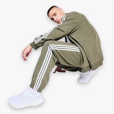 Mann in olivgrünem Trainingsanzug mit weißen Streifen, kniet auf dem Boden, trägt weiße Turnschuhe