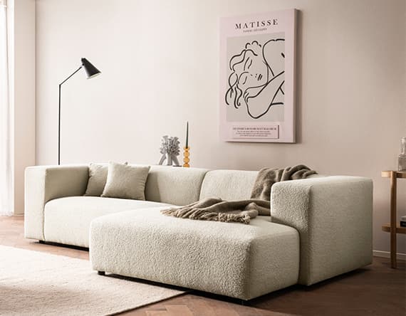 Gemütliches Wohnzimmer mit beigem Ecksofa, Decke, Kissen, Stehlampe und Matisse-Kunstdruck an der Wand