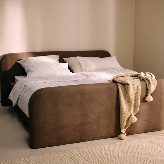 Großes Bett mit braunem Polsterrahmen, weißer Bettwäsche und beiger Strickdecke mit Quasten, steht in einem hellen Zimmer.