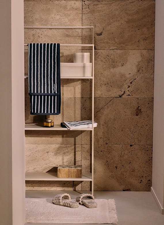 Weißes Regal mit blau-weiß gestreiften Handtüchern, Kerzen, Holzbox und Pantoffeln auf weißem Teppich vor einer Steinwand.