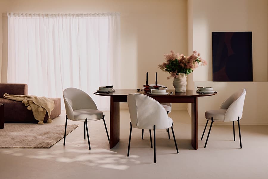 Esszimmer mit Holztisch, vier weißen Stühlen, gedeckt mit Geschirr, Vase mit Blumen, Kerzenständer, Sofa im Hintergrund, helles Fenster