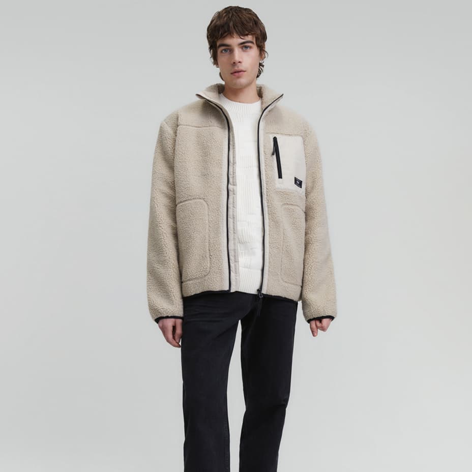 Mann trägt beige Fleecejacke mit Brusttasche, darunter weißer Pullover und schwarze Hose, Gesicht ist verschwommen