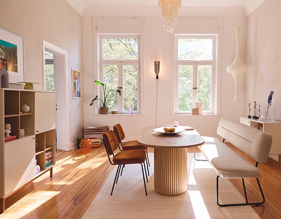 Esszimmer mit rundem Tisch, vier Stühlen und Bank, zwei Fenstern, Regal und Pflanzen, warmes Tageslicht füllt den Raum