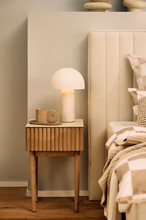 Nachtisch mit Lampe, Tasse und Untertasse neben einem Bett mit beige-weiß kariertem Bettzeug und beigem Kopfteil