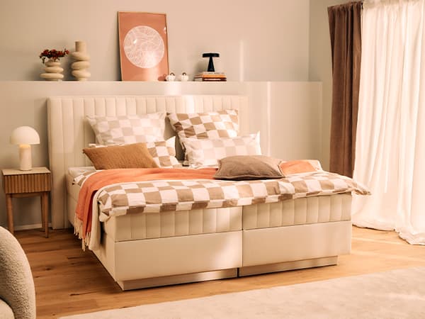 Hellbeiges Doppelbett mit karierten Kissen, orangefarbener Decke und Dekorationen auf dem Kopfteil im sonnigen Schlafzimmer.