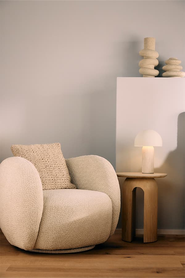 Beiger Sessel mit passendem Kissen neben einem Holztisch mit Lampe und dekorativen Objekten in einem modernen Raum
