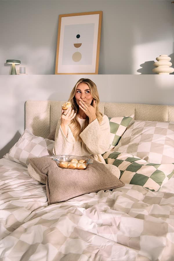 Frau sitzt auf einem Bett mit einer Schale Gebäck, trägt einen Bademantel, im Hintergrund ein gerahmtes Bild an der Wand