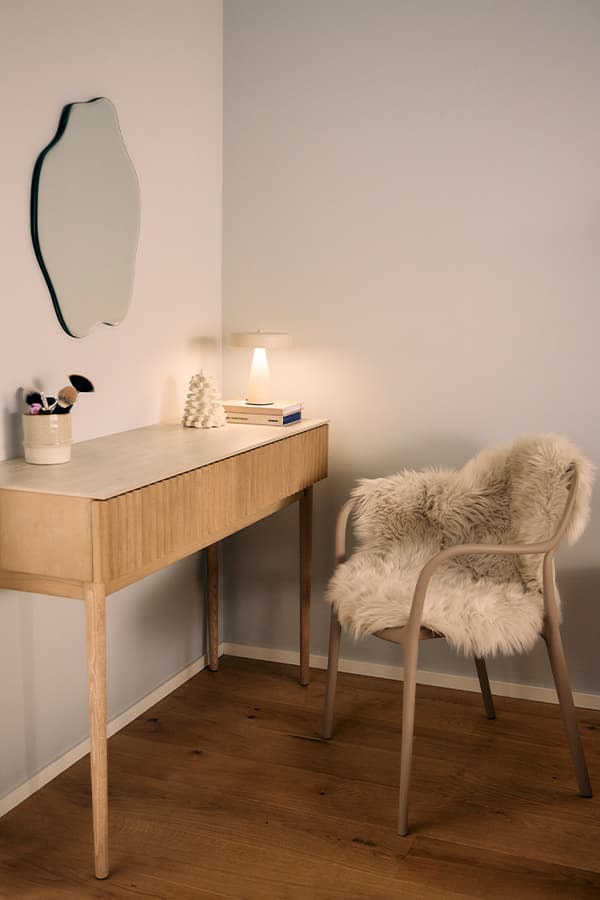Schlichter Schminktisch mit Spiegel und Lampe. Daneben ein Stuhl mit Fellauflage auf Holzfußboden.