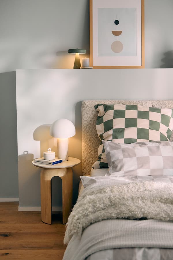 Nachttisch mit Lampe und Tasse neben Bett mit karierten Kissen, Wandregal mit Kerze und geometrischem Bild darüber