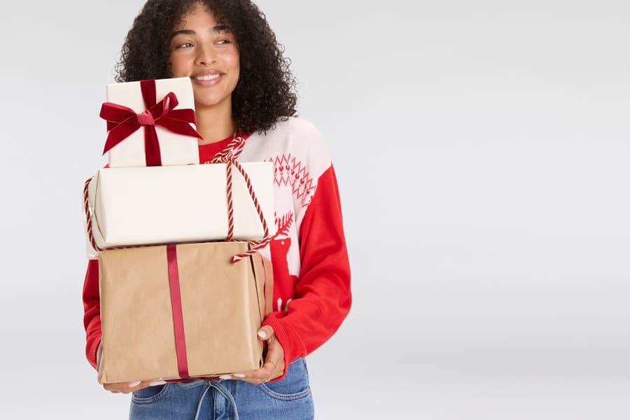Frau in rotem Weihnachtspullover trägt drei gestapelte Geschenke mit roten Bändern und Schleifen.