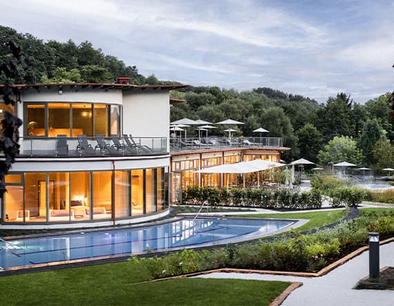 Modernes Wellnesshotel mit Pool und Terrasse, umgeben von grüner Natur und Bäumen, bei Sonnenuntergang
