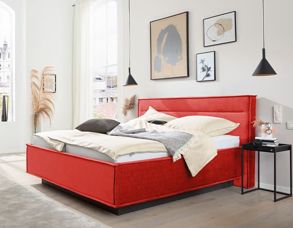 Modernes Schlafzimmer mit rotem Bett, beige Bettwäsche und schwarzem Nachttisch. Drei Bilder hängen an der Wand.