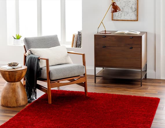 Sessel mit grauem Polster und Kissen, roter Teppich, Beistelltisch, Kommode mit Lampe und Buch in hellem Raum