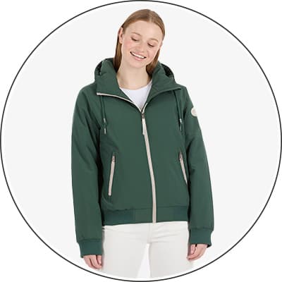 Frau trägt grüne Jacke mit Kapuze und Reißverschlüssen, darunter weißes Shirt und weiße Hose