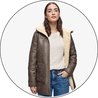 Frau trägt eine braune Winterjacke mit hellem Innenfutter, dazu einen cremefarbenen Pullover und blaue Jeans.