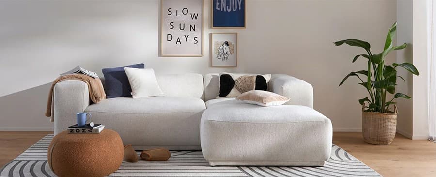 Slow Sundays – gemütliches Wohnzimmer mit weißem Sofa, großen Kissen, braunem Hocker und Pflanze im Korb auf gestreiftem Teppich